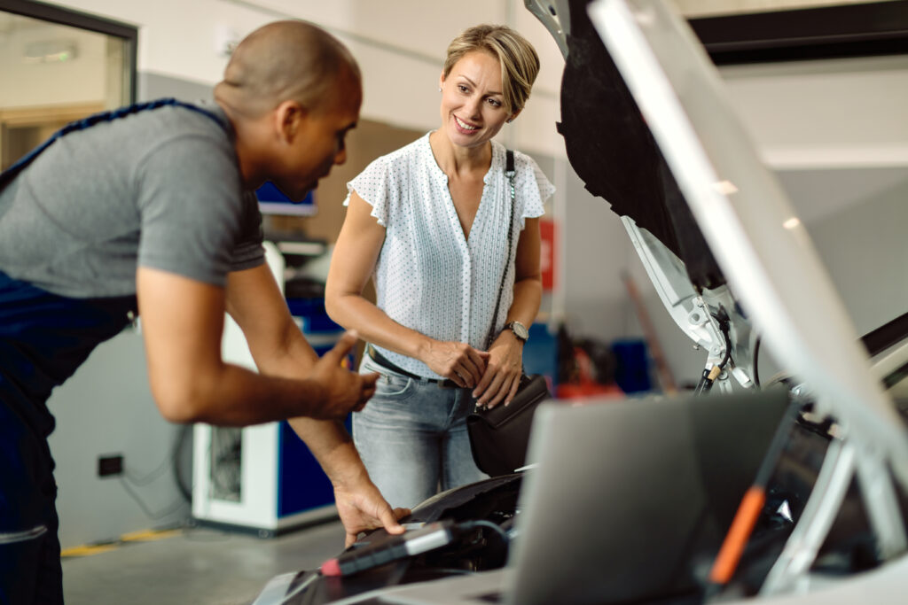 Mecánico explicando el funcionamiento del motor a una mujer en un taller de automóviles, mientras ella observa atentamente y sostiene un bolso.